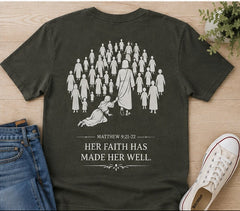 An olive green graphic t-shirt laid flat on a light wood background, styled with blue jeans, a white sneaker, and a small potted plant. The back of the t-shirt features a white line-art illustration of a crowd of people with one figure reaching down to a kneeling woman, accompanied by the text 'MATTHEW 9:21-22 HER FAITH HAS MADE HER WELL.